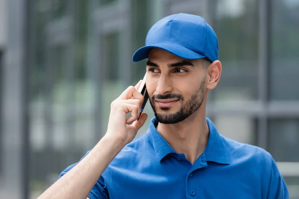 Mensajero árabe sonriente en gorra hablando en el teléfono inteligente al aire libre - foto de stock