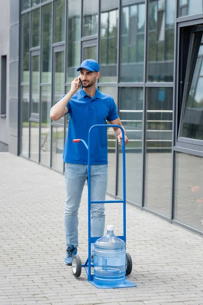 Arabian deliveryman in uniform talking on smartphone near bottle of water on cart and building — Stock Photo