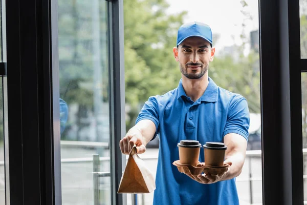 Mensajero musulmán sosteniendo vasos de papel y bolsa cerca de la puerta del edificio - foto de stock