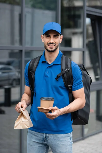 Alegre mensajero musulmán con mochila térmica, café para llevar y bolsa de papel mirando a la cámara al aire libre - foto de stock