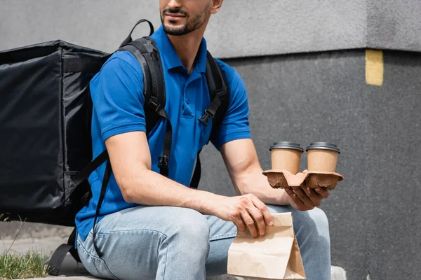 Vista cortada do correio com mochila térmica segurando saco de papel e copos takeaway ao ar livre — Fotografia de Stock