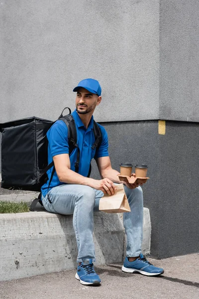 Young arabian deliveryman with thermo backpack holding paper cups and bag outdoors — Stock Photo