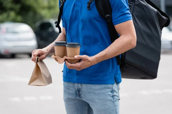 Cropped view of courier with thermo backpack holding paper cups and bag outdoors — Stock Photo