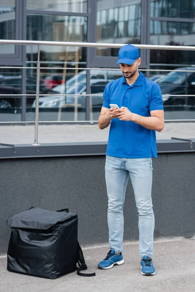 Muslim deliveryman in uniform using smartphone near thermo backpack outdoors — Stock Photo
