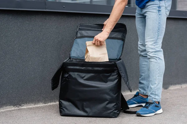 Vista recortada del repartidor tomando bolsa de papel de la mochila termo al aire libre - foto de stock