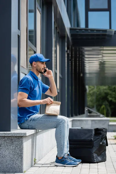 Vista laterale del fattorino musulmano che parla su smartphone e tiene il sacchetto di carta vicino allo zaino termico e all'edificio — Foto stock