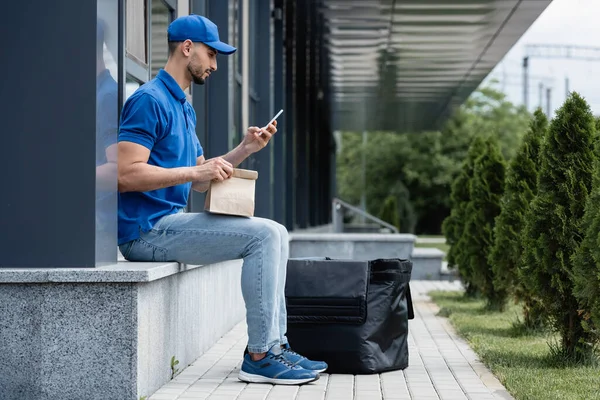 Vista lateral del repartidor musulmán con teléfono inteligente y bolsa de papel cerca de la mochila térmica al aire libre - foto de stock