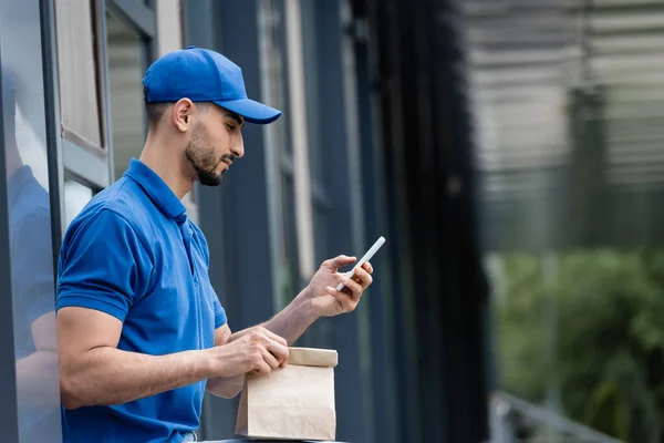 Side view of muslim courier in cap using smartphone and holding paper bag near building — Stock Photo