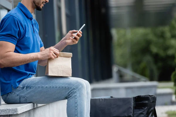 Vista cortada do correio sorridente usando smartphone e segurando saco de papel ao ar livre — Fotografia de Stock