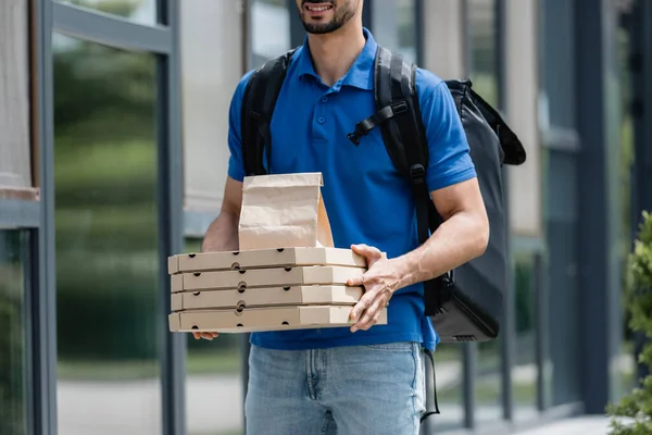 Vista recortada del repartidor alegre sosteniendo cajas de pizza y bolsa de papel cerca del edificio - foto de stock