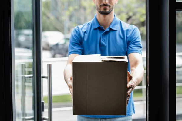 Vista recortada del mensajero borroso que sostiene la caja de cartón cerca de la puerta - foto de stock