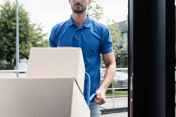 Cropped view of courier standing near boxes and cart outdoors — Stock Photo