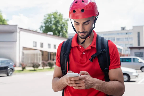 Arabian courier in helmet using smartphone on urban street — Stock Photo