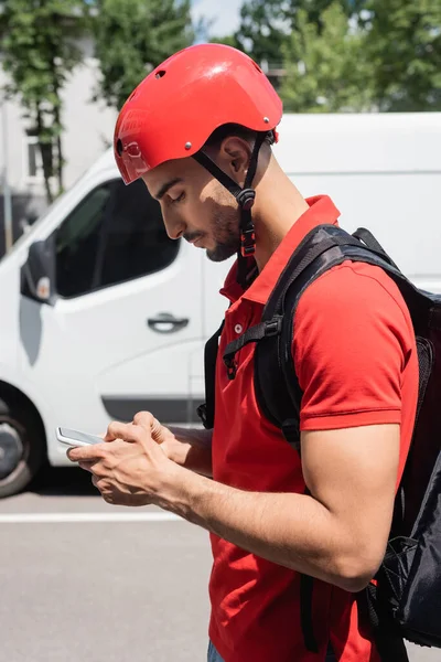 Vista laterale del corriere arabo in casco utilizzando smartphone su strada urbana — Foto stock