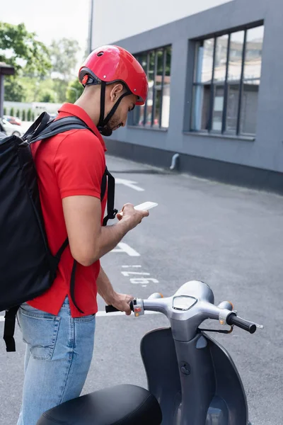 Arabian delivery en casco usando smartphone cerca de scooter al aire libre - foto de stock