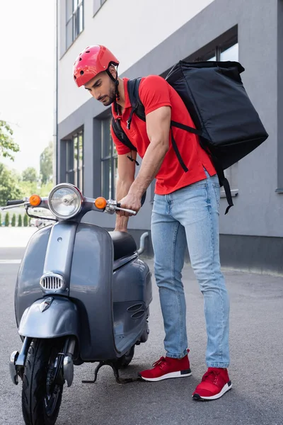 Correio árabe com mochila térmica em pé perto de scooter ao ar livre — Fotografia de Stock