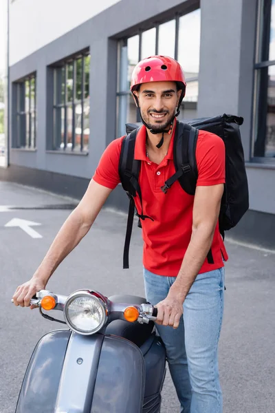 Mensajero musulmán sonriente con mochila térmica de pie cerca de scooter - foto de stock