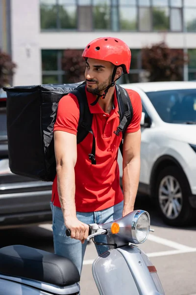 Arabian deliveryman in helmet and thermo backpack standing near scooter outdoors — Stock Photo