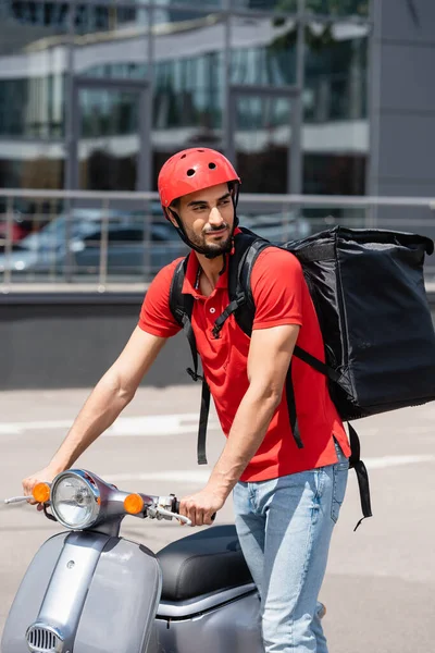 Arabian deliveryman in helmet and thermo backpack standing near scooter outdoors — Stock Photo