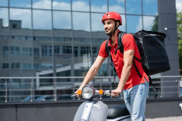 Livreur musulman souriant dans le casque et le sac à dos thermique regardant loin près de scooter à l'extérieur — Photo de stock