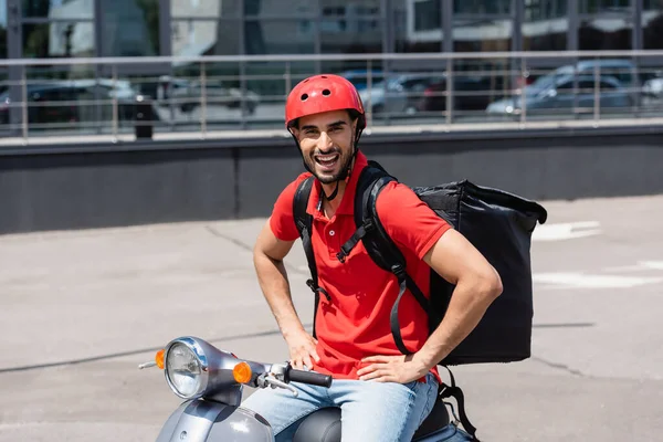 Emocionado repartidor árabe con mochila termo cogido de la mano en las caderas mientras está sentado en scooter — Stock Photo