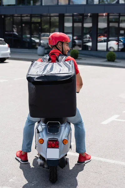 Seitenansicht eines lächelnden muslimischen Lieferanten mit Thermo-Rucksack, der auf einem Roller auf der städtischen Straße sitzt — Stockfoto