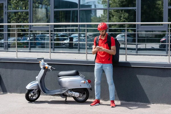 Young arabian deliveryman with thermo backpack using smartphone near scooter and building outdoors — Stock Photo