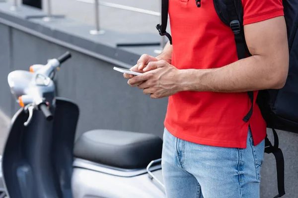 Vue recadrée du courrier avec sac à dos thermique à l'aide d'un smartphone près de scooter flou — Photo de stock