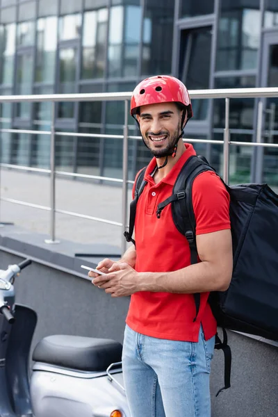 Repartidor árabe con mochila térmica y teléfono celular sonriendo a la cámara cerca de scooter borroso - foto de stock