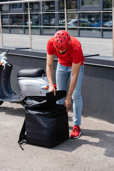 Joven mensajero en casco abriendo la mochila térmica cerca de scooter y edificio al aire libre — Stock Photo