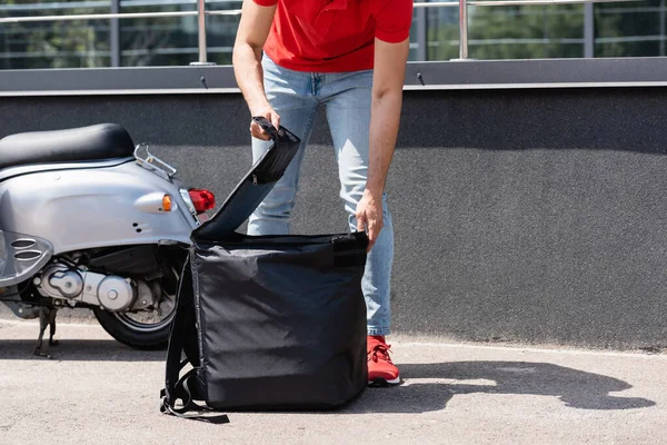 Cropped view of deliveryman opening thermo backpack near scooter — Stock Photo