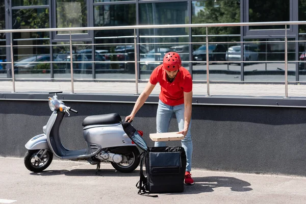 Entregador árabe segurando caixa de pizza perto de mochila térmica e scooter ao ar livre — Fotografia de Stock