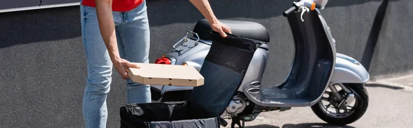 Vista recortada del repartidor sosteniendo la caja de pizza cerca de la mochila termo abierta y el scooter al aire libre, pancarta - foto de stock