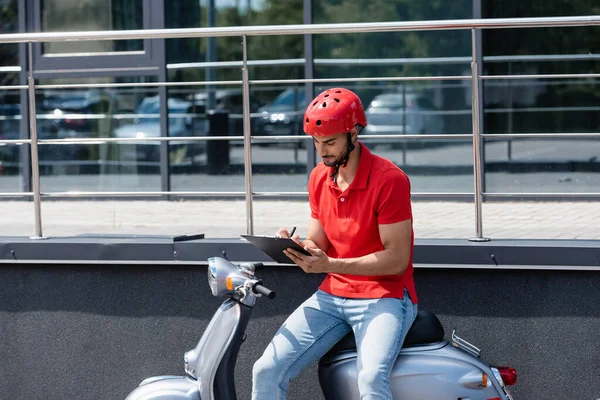 Junger muslimischer Kurier mit Helm schreibt auf Klemmbrett, während er auf Roller im Freien sitzt — Stockfoto