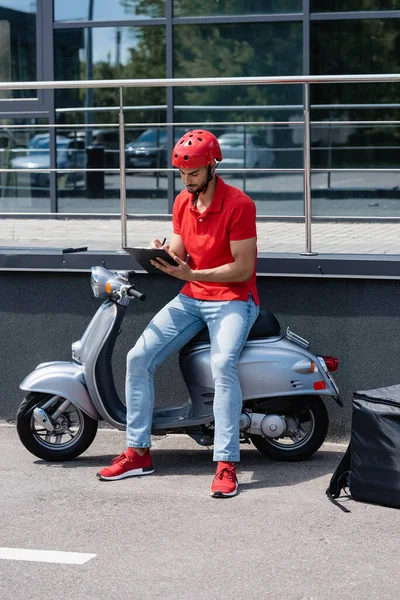 Young muslim deliveryman writing on clipboard on scooter near thermo backpack outdoors — Stock Photo