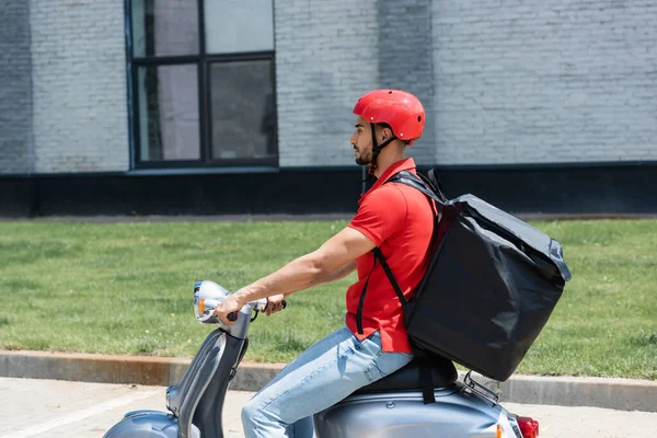 Vista laterale del corriere musulmano con termo zaino in sella scooter su strada urbana — Foto stock