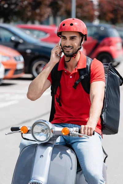 Smiling arabian deliveryman with thermo backpack talking on smartphone while riding scooter — Stock Photo
