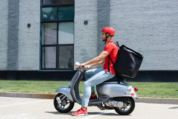 Vista lateral do correio muçulmano em uniforme vermelho e mochila térmica montando scooter ao ar livre — Fotografia de Stock