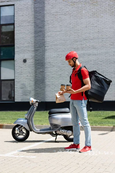 Vista lateral del mensajero árabe con mochila térmica sosteniendo café para llevar y bolsa de papel cerca de scooter - foto de stock