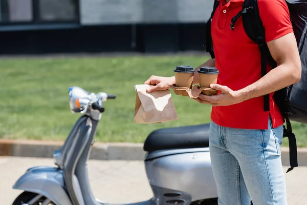 Vue recadrée du livreur avec sac à dos thermique tenant des tasses en papier et sac près de scooter flou — Photo de stock