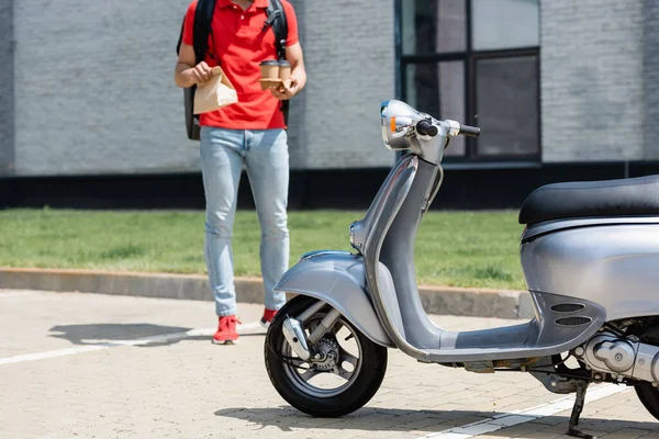 Cropped view of scooter near blurred courier with coffee to go and paper bag outdoors — Stock Photo