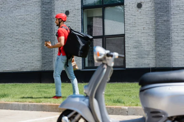 Smiling arabian deliveryman with coffee to go and paper bag walking on grass near blurred scooter — Stock Photo