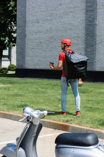 Vue latérale du livreur musulman positif avec sac en papier et tasses marchant sur l'herbe près du scooter flou — Photo de stock