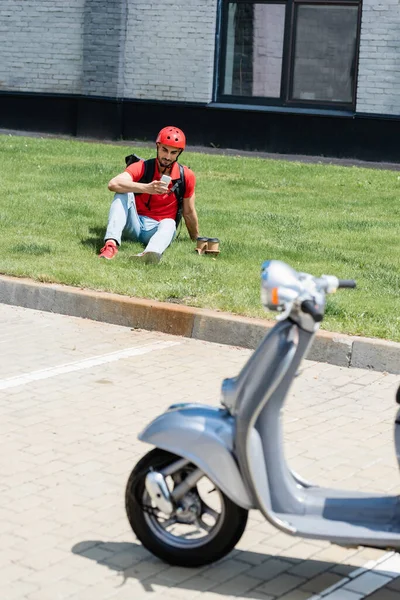 Arabian courier using smartphone near coffee to go on grass and blurred scooter — Stock Photo