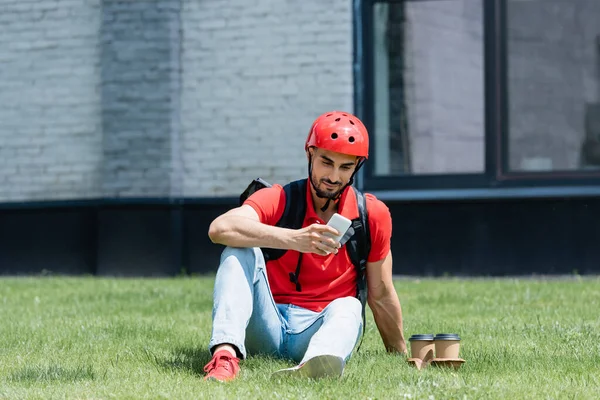 Courrier arabe avec sac à dos thermique utilisant un smartphone sur l'herbe — Photo de stock