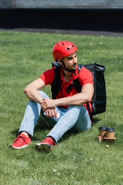 Junger muslimischer Kurier mit Helm und Thermo-Rucksack hält Smartphone in der Nähe von Pappbechern auf Gras — Stockfoto