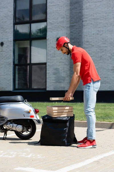 Seitenansicht des arabischen Kuriers im Helm, der Pizzakartons in der Nähe von Thermo-Rucksack und Roller hält — Stockfoto