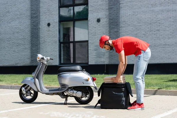 Vue latérale du courrier arabe dans un casque prenant des boîtes à pizza du sac à dos thermique près du scooter à l'extérieur — Photo de stock