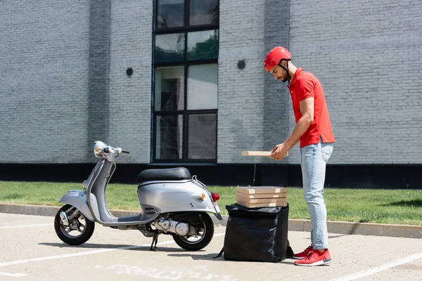 Vista lateral do jovem muçulmano entregador no capacete segurando caixa de pizza perto de mochila térmica e scooter — Fotografia de Stock