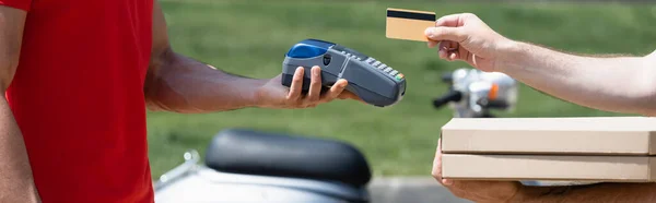 Vista recortada del hombre con cajas de pizza con tarjeta de crédito cerca de mensajero con terminal de pago al aire libre, pancarta - foto de stock